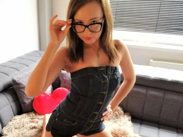 GeileJaqueline-hot in a denim strapless dress, black glasses, and a sexy smile, with a cute heart pillow on a gray couch, hand on hip, slightly bending forward.