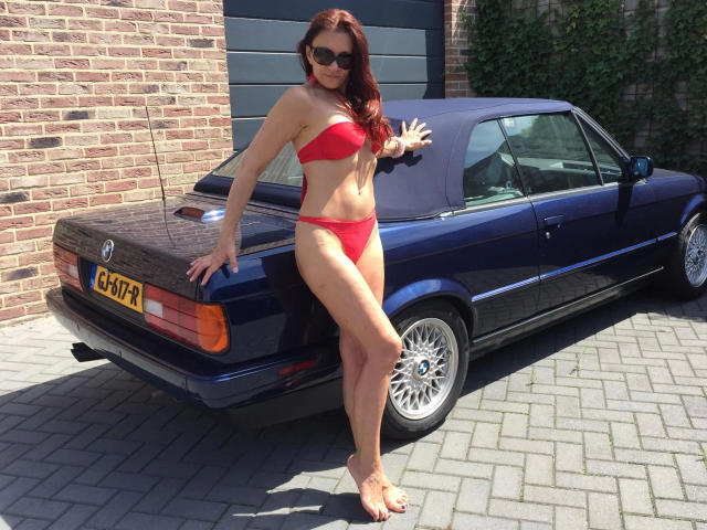 AnnaBronx-hot stands in a red bikini, showing her tits and ass, with a blue car, wearing sunglasses, touching the car's roof, and showing a tattoo on her ankle.
