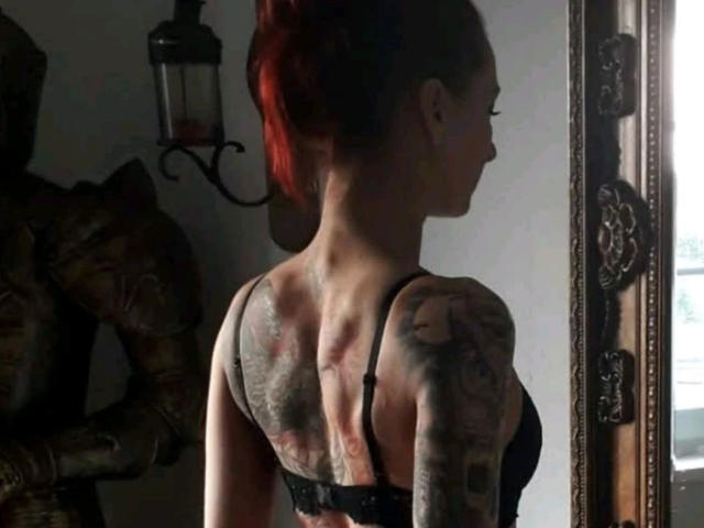 JennaLaCroix-hot with red hair in a bun, back tattooed, wearing black bra, standing in dimly lit room, side profile, muscular arms, and a dark lantern on wall.