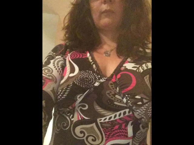 GeilesLuder-hot with curly brown hair, wearing a colorful, patterned dress with pink, black, and white swirls, and a necklace, has visible tits and a slight pout.