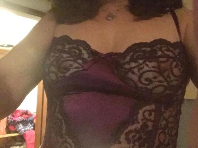 GeilesLuder-hot in a purple lace and satin corset, showing her tits, with black lace cups and a deep purple satin panel, wearing a heart necklace, in a dim room.