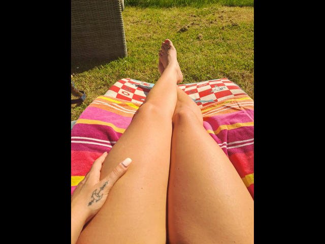 Le gambe di LinaaPuschel-hot sono distese su un asciugamano colorato a righe, con una mano che mostra un tatuaggio, e i suoi piedi puntano verso l'erba verde. La luce del sole mette in risalto la sua pelle abbronzata..