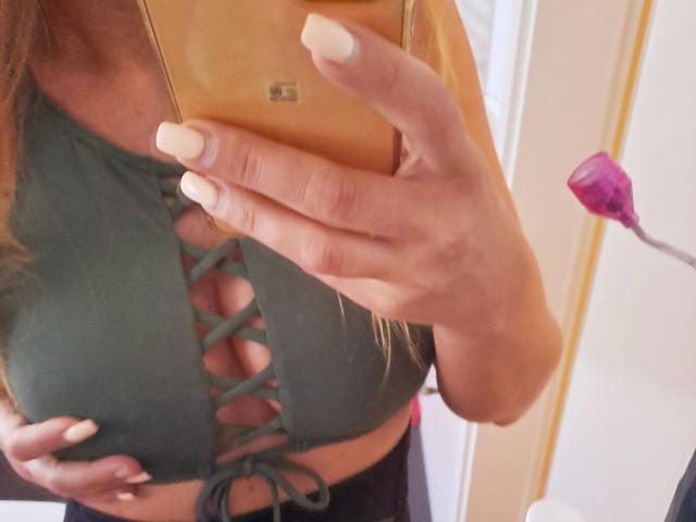LinaaPuschel-hot holding a phone in front of her tits, wearing a green lace-up crop top, showing off her white-manicured nails and tan skin.