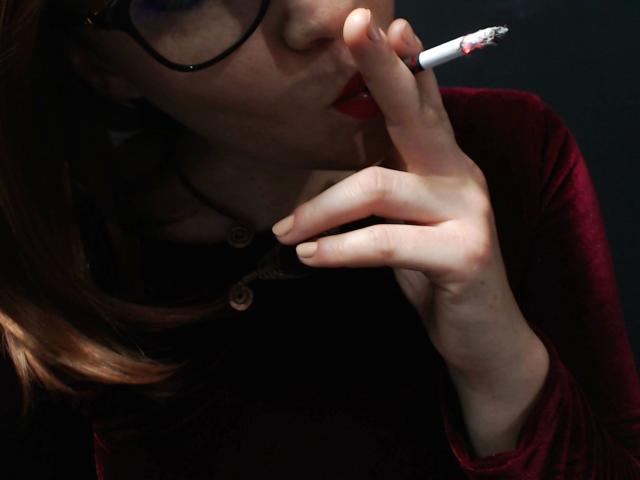 ScorpionLady-hot with red lips, holding a lit cigarette between her fingers, wearing glasses and a dark red velvet long-sleeve top, her hand near her face.