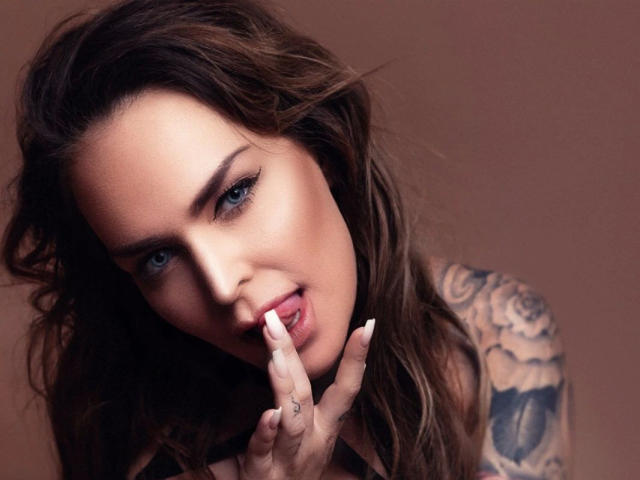 PenelopeLopez-hot with wavy brown hair, tattoos on her right arm, blue eyes, and pink lipstick, sensually biting her long, white-painted fingernail.