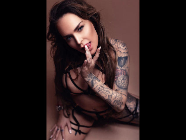 PenelopeLopez-hot with tattoos, brown wavy hair, and blue eyes, sucking on a finger, wearing black strappy lingerie, showing her tits and ass. She's kneeling, looking seductive.