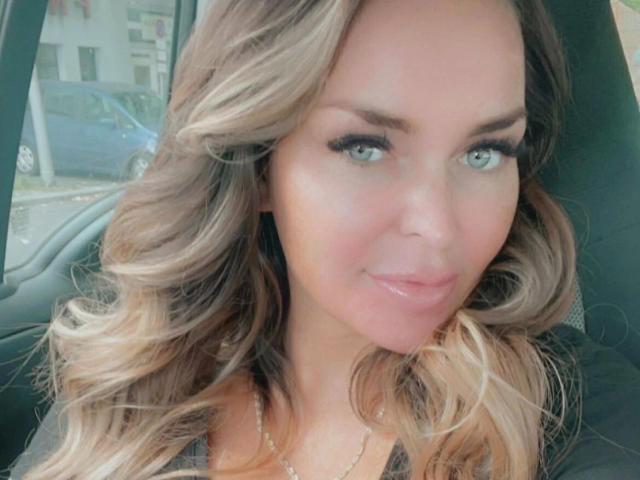 PenelopeLopez-hot has blonde, wavy hair, blue eyes, and glossy pink lips, sitting in a car with a subtle smile, wearing a black top. Her tits are slightly visible.