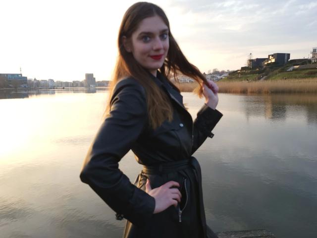 Zara-Bizarr with long brown hair, black leather jacket, and red lipstick, holding her hair, standing by a calm river, hands on hips, smiling, sunset lighting.
