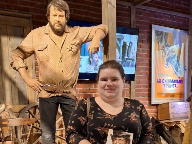 Bbwnicole-hot, chubby with brown hair in a black floral top, sits in front of a wooden, cowboy statue with a belt, standing, wearing a dirty shirt and jeans.