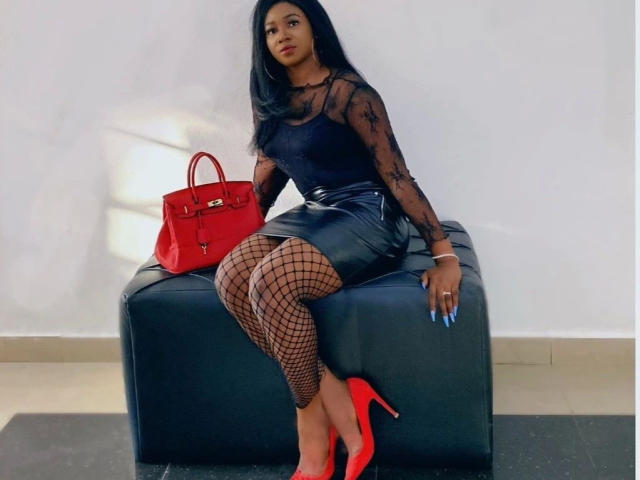 Goddessqueen-hot with black mesh top, black leather skirt, fishnet stockings, red high heels, and red purse, sitting on black ottoman, long black hair, blue nails.
