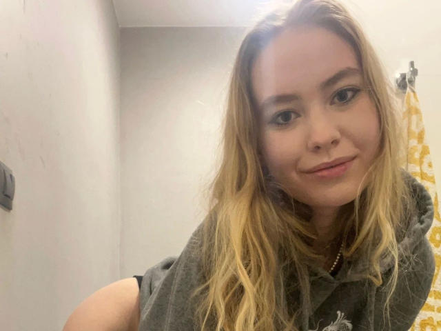 Blond BrandyHelga-hot with long hair, light skin, and subtle makeup, wearing a gray shirt, smiling slightly, and showing a nose ring in a bathroom selfie.