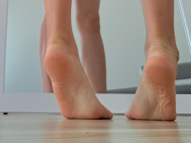 SourrSanndy-hot's soles are up, showing soft pink toes with slight wrinkles on wooden floor, reflected in a mirror. Bare legs, slightly visible, standing on toes.