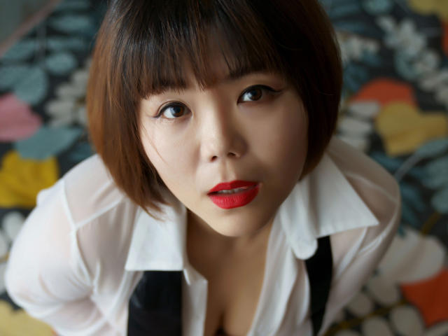 Laruina-hot with short brown hair, red lipstick, and white shirt, looking up with big brown eyes, showing cleavage, on colorful patterned bed.