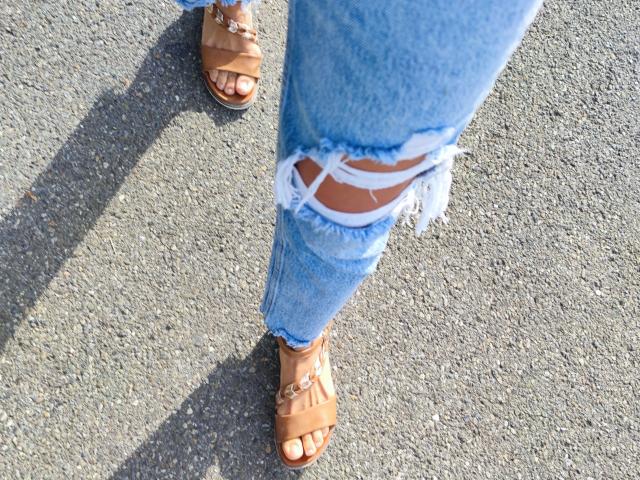 Lilita-Lush's ripped blue jeans with white straps, tan sandals with straps, and visible tan toes standing on a textured gray asphalt.
