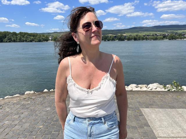 Pamm2023: una donna con la pelle abbronzata, capelli ricci scuri, che indossa occhiali da sole, un top bianco a spalline che mette in mostra piccoli seni, e jeans blu, in piedi vicino a un lago con una riva rocciosa..