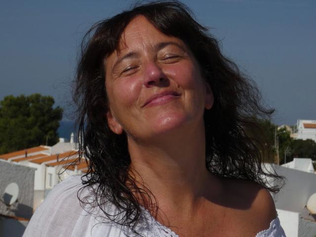 Pamm2023-hot, con gli occhi chiusi, sorridendo e con leggere striature sulla pelle dovute all'abbronzatura, indossa una maglietta bianca con una spalla scoperta, ha capelli neri, ondulati e umidi, e un viso leggermente lucido a causa del sudore..