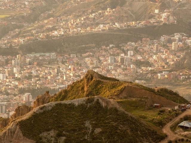 A sunlit mountain with green shrubs and rocky peaks in the foreground, overlooking a sprawling city with dense, multistory buildings and winding roads. InkedMandy-hot's ass is in the city.