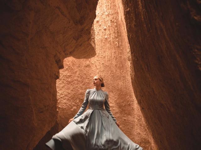 InkedMandy-hot stands in a rocky canyon, wearing a blue, long-sleeved dress, looking upward with a serene expression, surrounded by tall, brown rock walls.