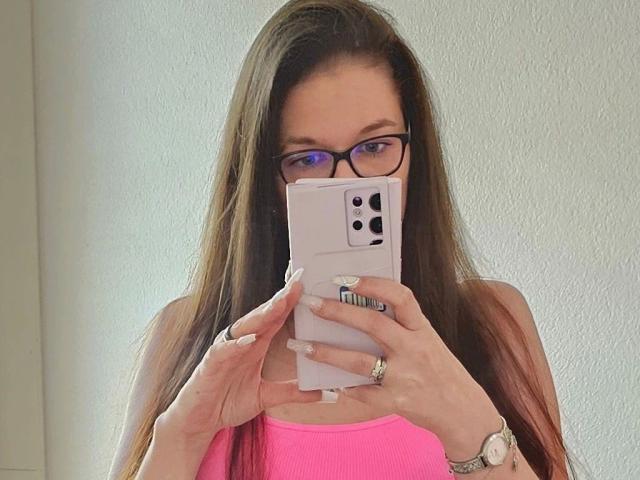 Zelda-hot with long brown hair, wearing glasses and a pink top, holding a pink iPhone, covering her tits with her hand, showing a ring and a watch.