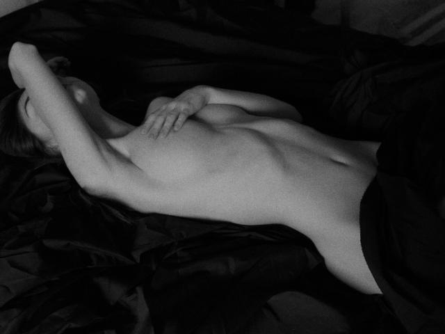 Fotografia in bianco e nero di una donna distesa su lenzuola scure, con le braccia sopra la testa, che copre i piccoli seni con una mano, ha l'addome piatto e il sedere leggermente visibile, in bianco e nero, con ombre delicate, in posizione rilassata..