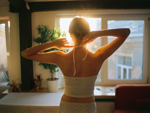 Barbell-hot with arms up, back to camera, sunlit, white strapless top, white polka dot panties, light brown hair in bun, window behind, potted plant to left.