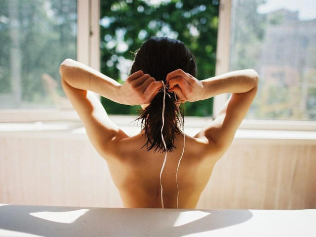 Ragazza con bilanciere, schiena rivolta verso la telecamera, capelli castani bagnati, braccia alzate, che si toglie le cuffie, filo bianco, luce solare, spalle scoperte, luce soffusa, interni, finestra sullo sfondo..