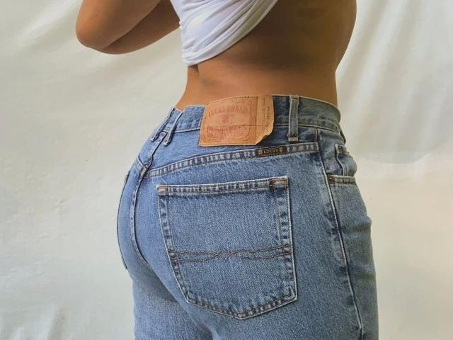 Das Mädchen mit dem Hintern, der durch die engen, blauen Jeans betont wird, trägt einen braunen Leder-Patch auf dem Rücken, der sichtbare Taschen und eine leichte Wölbung in der Taille zeigt. Sie hält ein weißes Hemd..