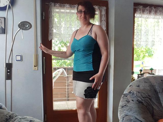EinfachDani-hot stands in a doorway, wearing a teal lace top, black and white skirt, and glasses, with a slightly curvy, average-ass physique, and short curly red hair.