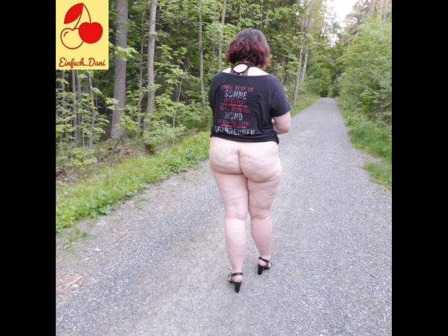 EinfachDani-hot has a curvy, pale-skinned body with a large ass, wearing a black shirt and black heels, walking on a gravel path with a forest background.