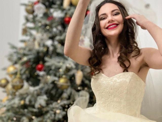 HairyMommy-hot with curly brown hair, red lipstick, smiling, hands up, wearing a strapless, lace, yellow dress, standing in front of a decorated Christmas tree.