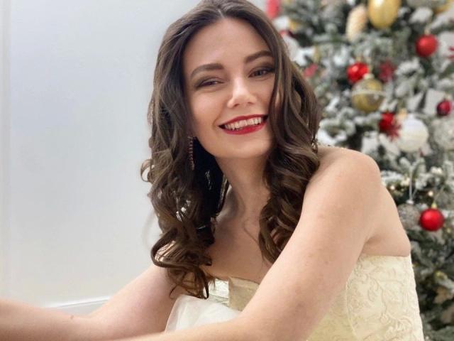 HairyMommy-hot with wavy brown hair, red lipstick, and a smile, wearing a white lace dress, sitting in front of a decorated Christmas tree.