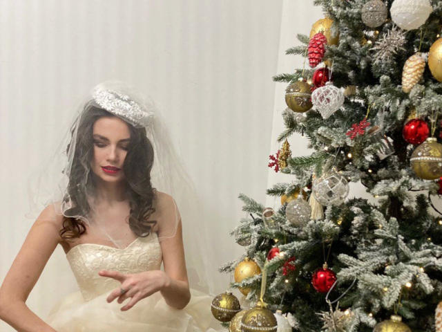 HairyMommy-hot with wavy brown hair, white bridal dress, and veil, looking down, standing next to a decorated Christmas tree with red, gold, and silver ornaments.
