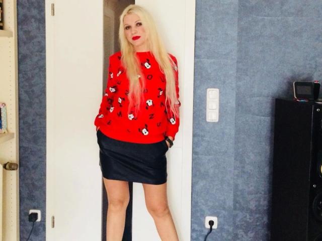 Sexy-hot woman with long blonde hair, red sweater with black skull patterns, black mini skirt, hands in pockets, standing in doorway, wearing red lipstick, blue walls.