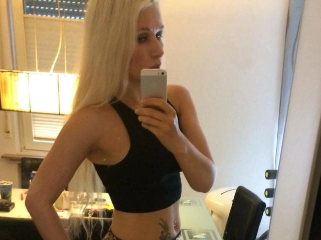 A blonde sexicandy-hot woman with small tits, wearing a black sports bra, takes a mirror selfie, showing a tattoo on her right side. She holds a silver phone.