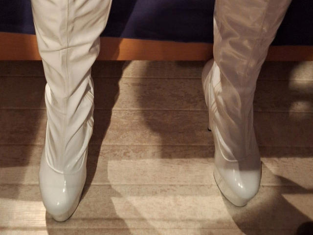 Bootsica-hot's white, cuffed, knee-high boots with a wavy design and shiny, pointed-toe heels standing on wooden floor, shadowed from above.