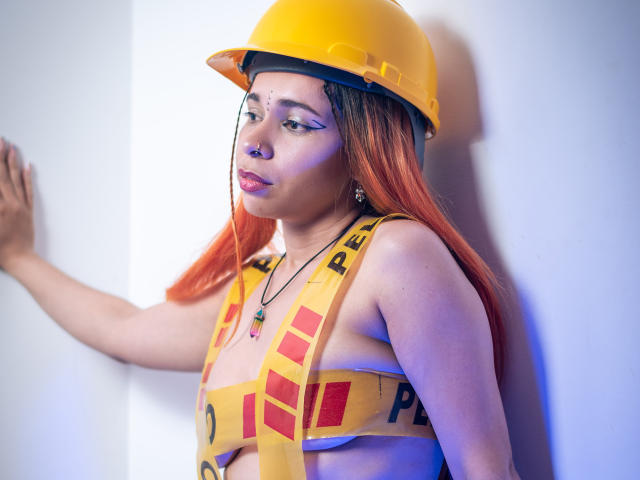 MarthaMaya with orange hair, yellow helmet, and yellow "PE" safety vest, exposing her tits, standing against a white wall, looking to the side with a pouty expression.