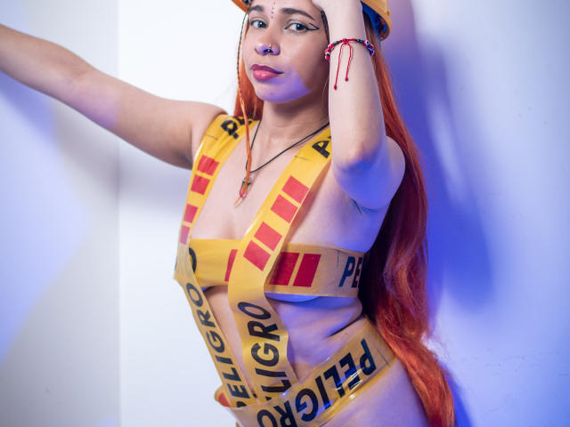 MarthaMaya with long red hair, wearing yellow caution tape covering her tits and belly, arm raised, hand on head, blue lighting, red bracelet, and a confident expression.