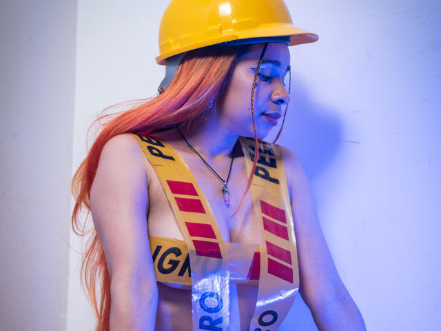MarthaMaya with orange hair, yellow helmet, and yellow reflective vest, exposing her tits. She's looking down, with blue lighting. Necklace with cross. Sexy construction worker vibe.