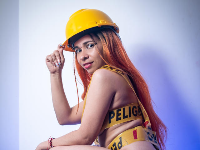 MarthaMaya with orange hair, yellow helmet, and yellow "PELIGRA" tape bikini, showing her tits, sitting, holding helmet, blue light, red bracelet, smiling, looking at camera.