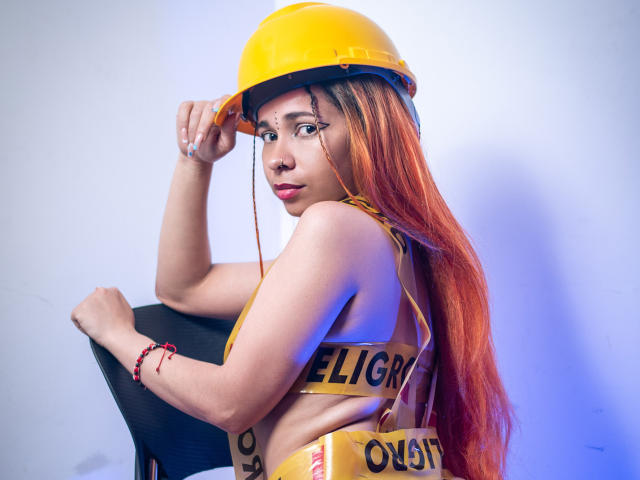 MarthaMaya with orange hair, wearing yellow helmet and "OIL" strap, looking over shoulder, sitting on black chair, showing side-boob, blue light, red bracelet.