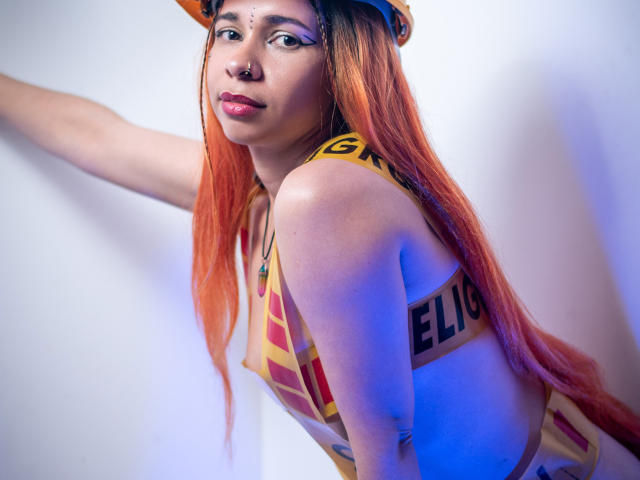 MarthaMaya mit orangefarbenem Haar, die ein gelb-rotes Streifenoberteil trägt, hat eine sichtbare Brust, einen Nasenring und ein Gesichtspiercing, lehnt sich an eine weiße Wand und schaut in die Kamera..