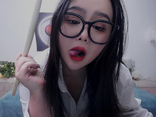 TinaFany with black glasses, tongue out, holding a paddle, wearing a white shirt, has long black hair, red lipstick, and a nose piercing, looking seductive.