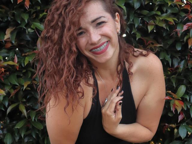 ScarlettthVega has curly red hair, smiling with glittery blue teeth, wearing a black tank top, touching her chest with a ringed hand, against a leafy background.