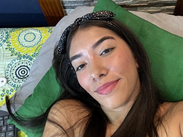 SharonKlart with black hair, wearing a polka dot headband, lying on a green pillow, smiling with pink lips, fair skin, and dark eyebrows, looking relaxed. Boobs not visible.