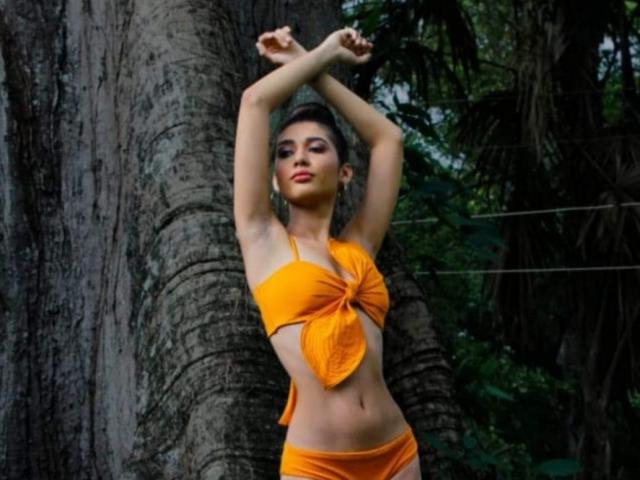 PauletteRoy stands against a tree with her hand on her head, wearing an orange bikini, showing her tits and ass, with dark roots and green leaves.