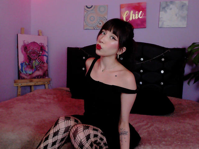 IzzysMisss with black hair, red lipstick, and a black tank top, touching her cheek with a hand, showing a small heart tattoo on her chest.