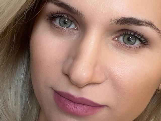 DariaBloom's face, with her green eyes, blonde hair, and pink lipstick, showing a subtle smile, has smooth skin and neatly shaped eyebrows.