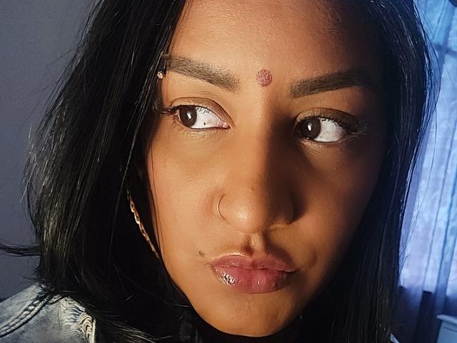 Nahirr has dark brown skin, black straight hair, and a small red dot on her forehead. She's looking to the side with glossy pink lips and a nose ring.