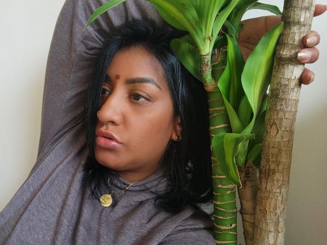 Nahirr with dark hair, brown skin, and a red dot on her forehead, holding a plant against a white wall, wearing a gray long-sleeve shirt. Nahirr's face is relaxed.