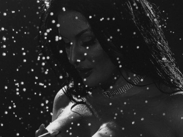Black and white photo of ClaireWarner with wet hair, sparkling necklace, and large bare breasts, eyes closed, surrounded by bokeh light dots, looking sexy and seductive.